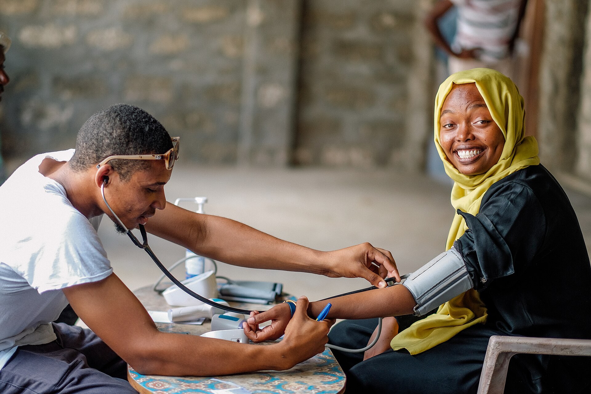 A health worker with a stethoscope takes a smiling woman's blood pressure at a table during a Safari Doctors clinic in Lamu County.