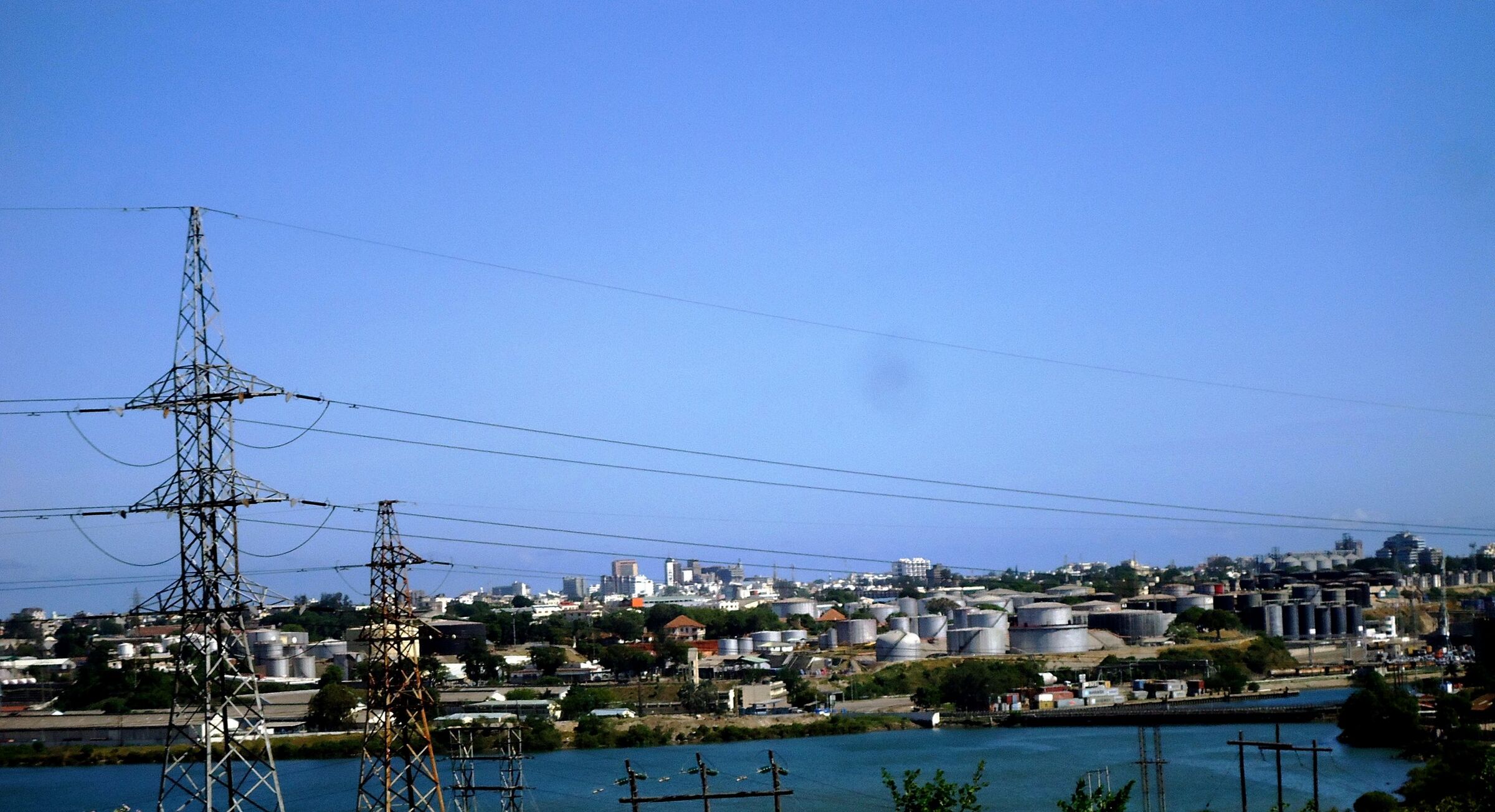 Rows of white industrial storage tanks along the Mombasa waterfront, seen across the water with electricity pylons in front and city buildings behind.