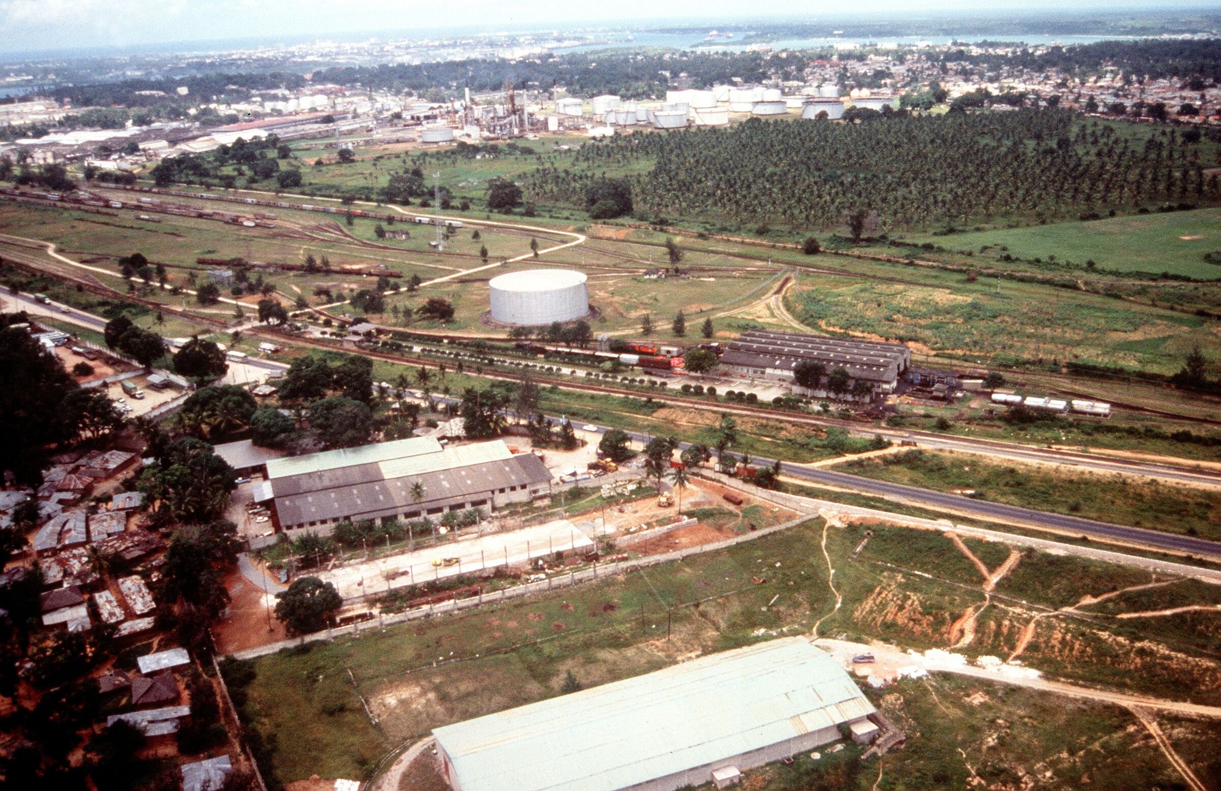 Aerial photograph from 1994 of Mombasa's mainland industrial area near the airport, with factory and warehouse roofs in the foreground, a railway yard and a large storage tank in the middle distance, and refinery tanks and a coconut plantation beyond. It shows the general area, not a project site.