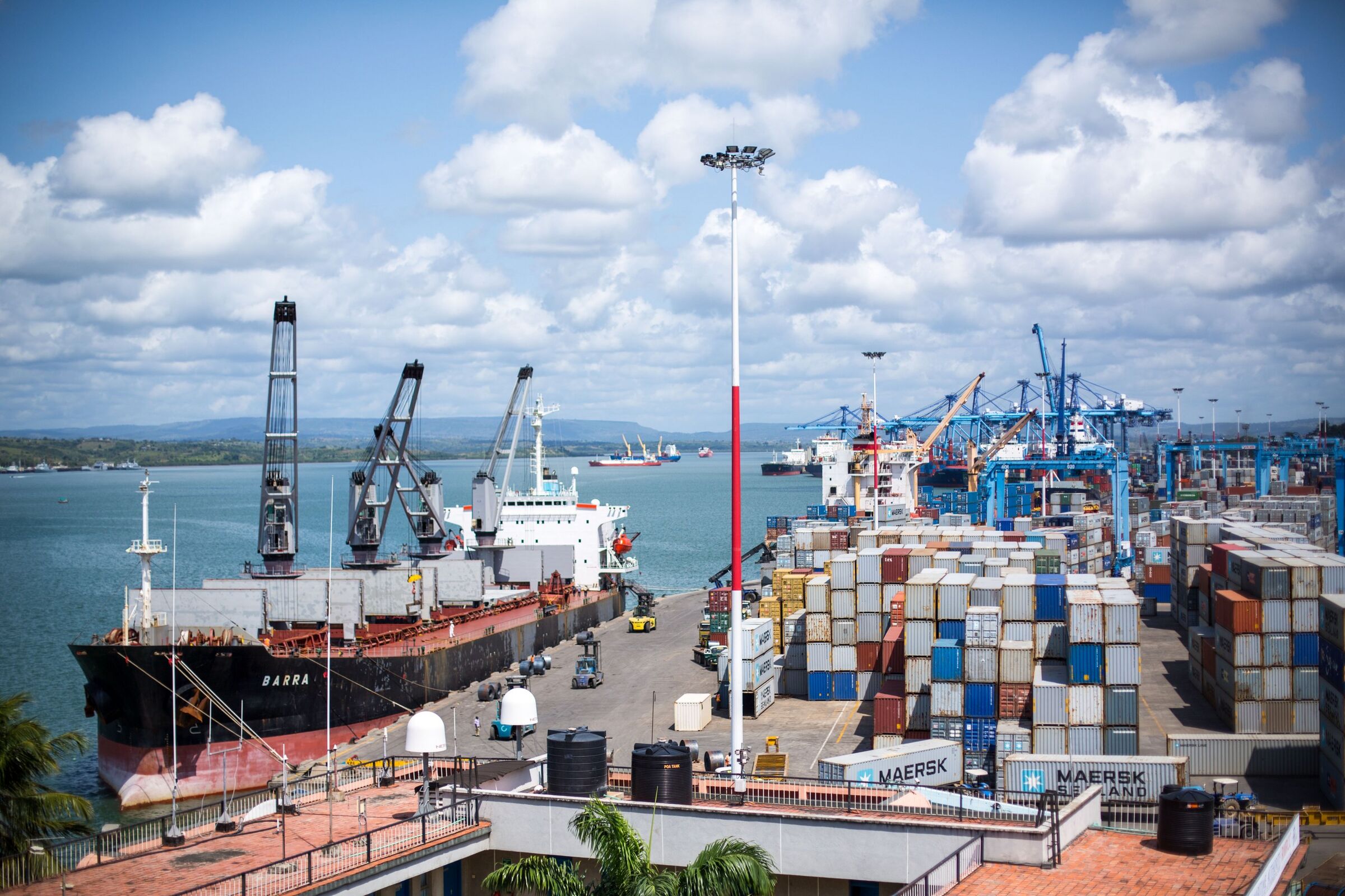 The Port of Mombasa in 2015: a bulk carrier at the quay in the foreground, stacked containers and blue container gantry cranes behind, and ships at anchor in the harbour