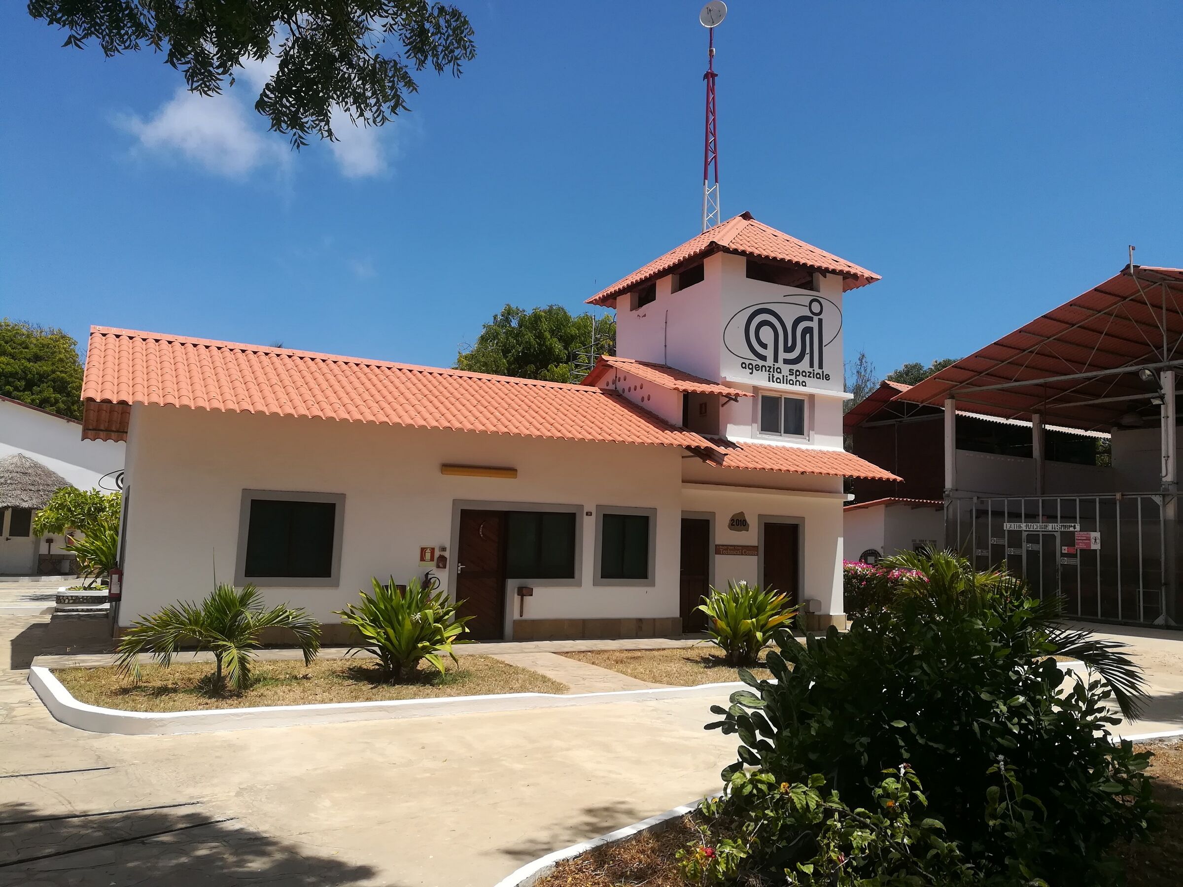 The Italian Space Agency's building at the Luigi Broglio Space Centre in Malindi, a white single-storey building with a red tiled roof and a small tower, under a blue sky. It is an existing facility, not the site of the proposed university.